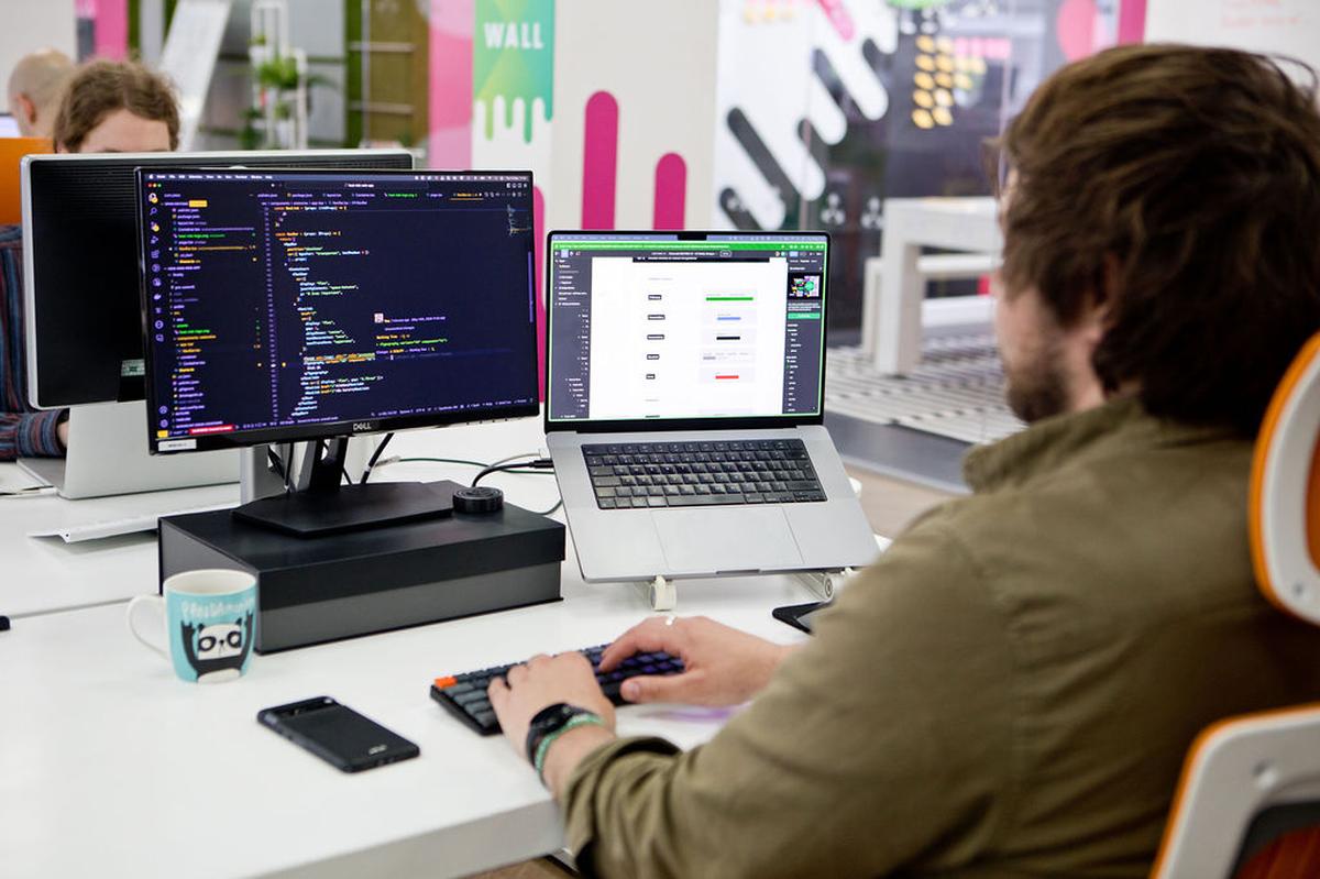 A person at a desk is programming on a dual monitor setup, featuring code on one screen and a web interface on the laptop, in an office environment.