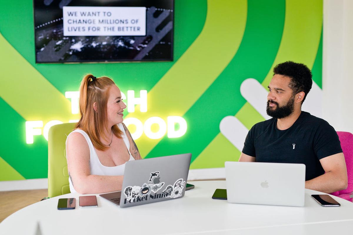 Two people sit at a table with laptops, discussing in a bright office with a green "Tech for Good" wall and motivational text on a screen.