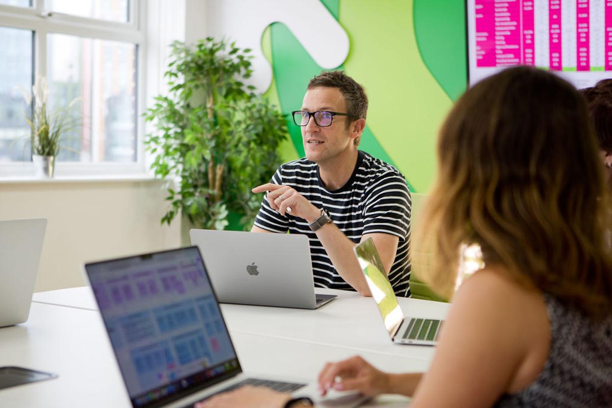 A man in glasses and a striped shirt speaks during a meeting, surrounded by colleagues with laptops in a bright office.