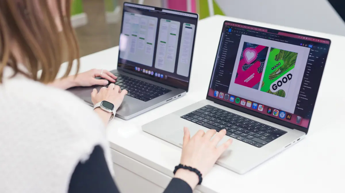 Two people working on laptops at a white desk; one screen shows design software, the other a workflow diagram.