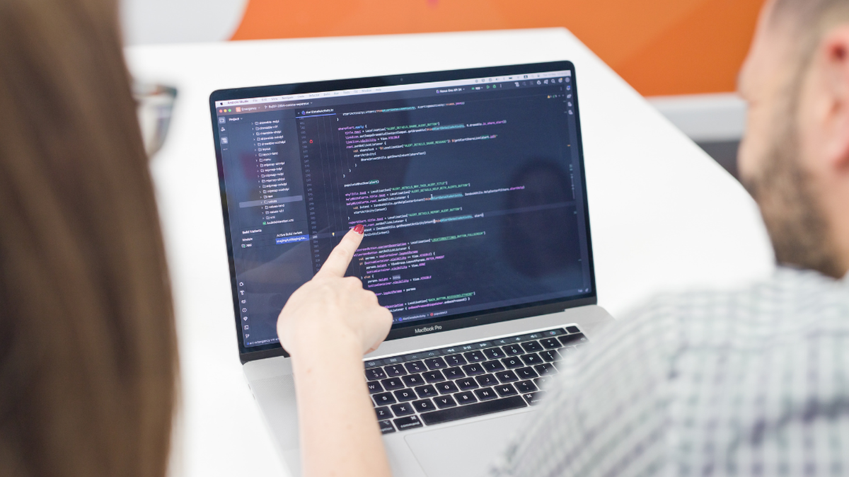 Two people looking at a MacBook screen displaying a code editor, with one person pointing at the code.