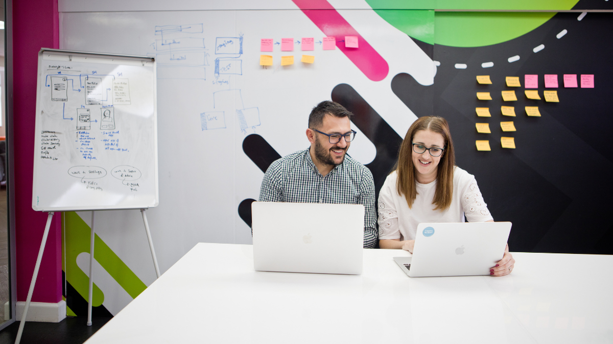 Two colleagues collaborate at a table with laptops, whiteboard diagrams, and sticky notes in a modern office setting.