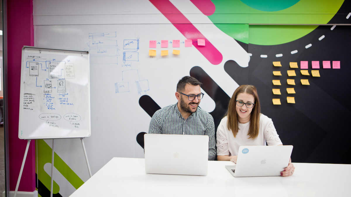 Two people working on laptops in a colorful office, with whiteboard diagrams and sticky notes on the wall behind them.