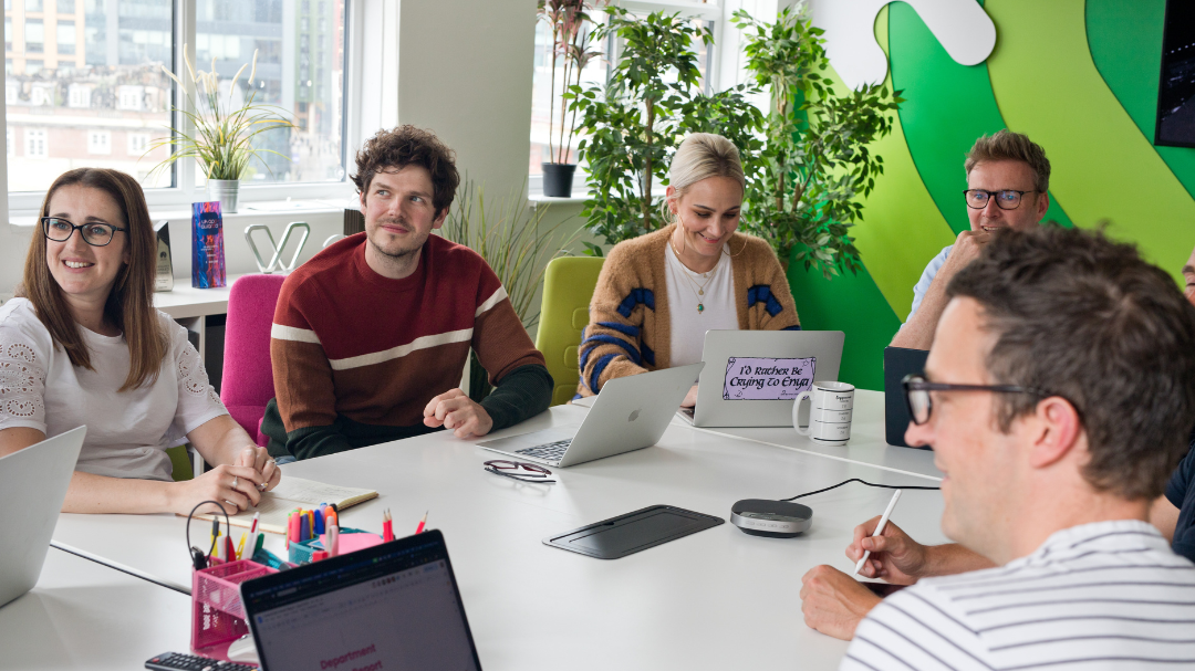 Group of people engaged in a meeting around a big table with laptops, notebooks, and plants in the background.