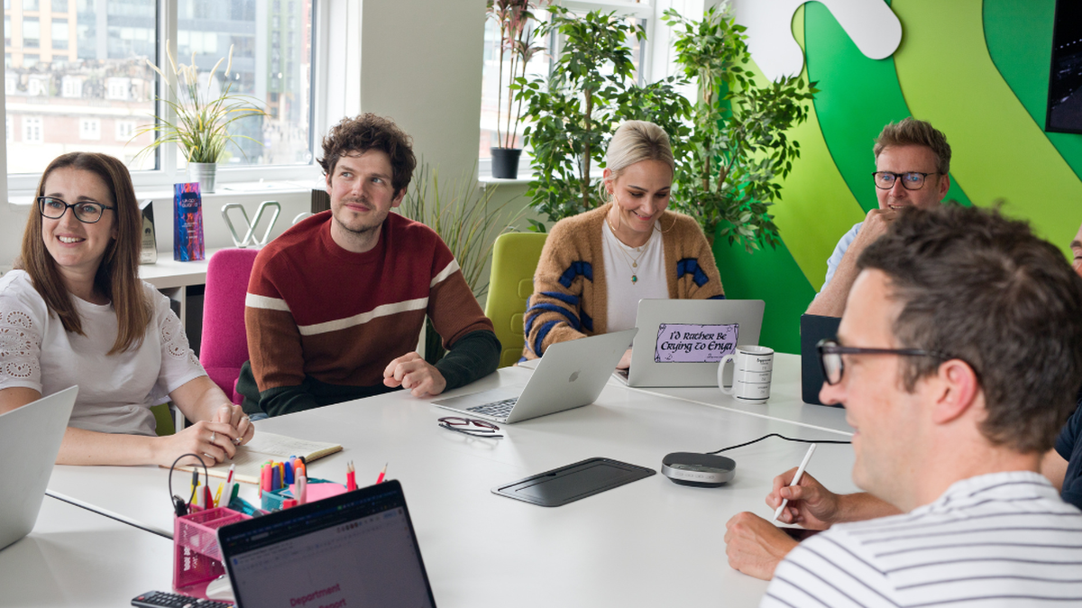 Group of people engaged in a meeting around a big table with laptops, notebooks, and plants in the background.