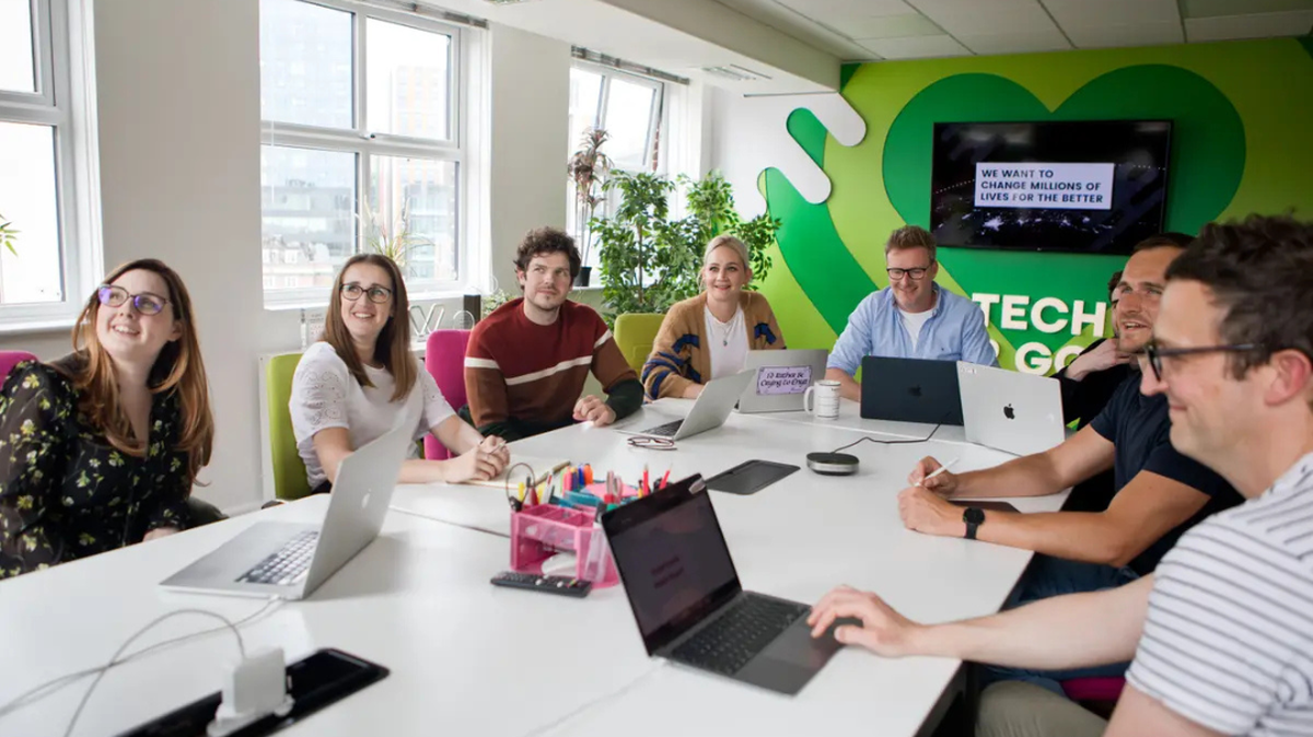 A group of people smiling and talking around a table with laptops in a bright office.