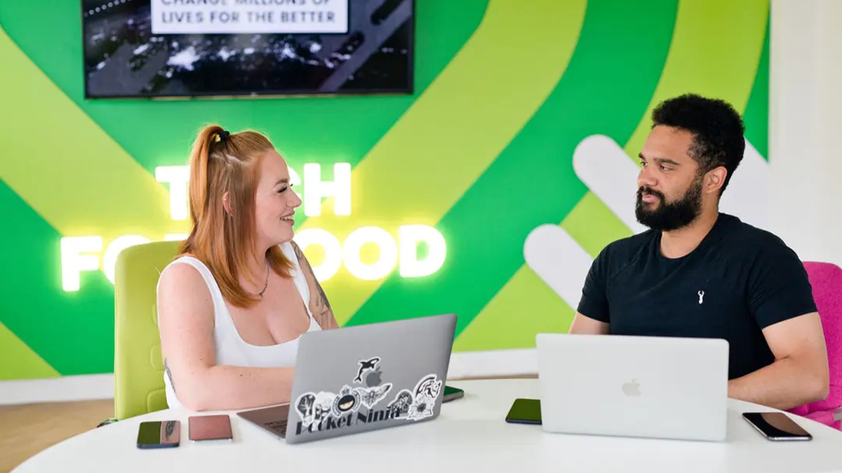 Two people sit at a table with laptops, chatting. A bright green wall with text forms the background.