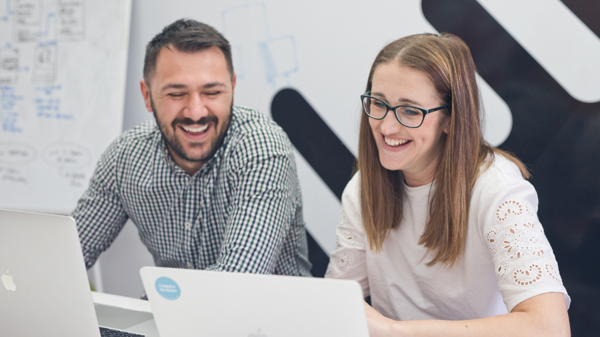 Two people smiling while looking at laptops, sitting at a desk with a whiteboard in the background.
