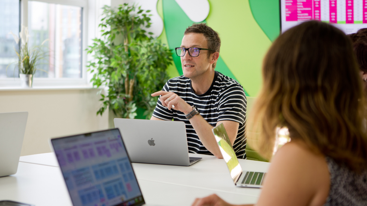 A man in glasses and a striped shirt speaks during a meeting, surrounded by colleagues with laptops in a bright office.