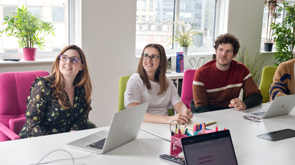 Three people sitting at a conference table with laptops and notes, attentively listening in a bright, modern office.