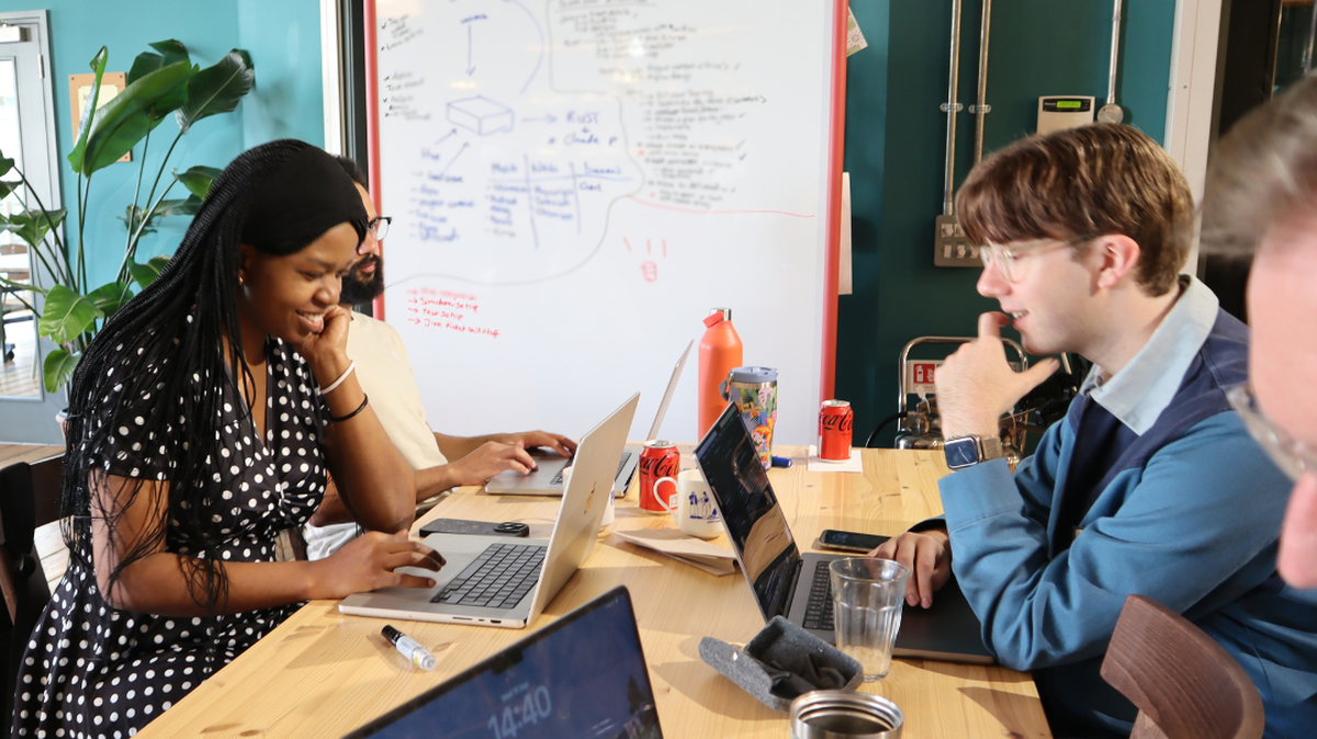 A group of people at a table working on laptops with a whiteboard covered in notes in the background.