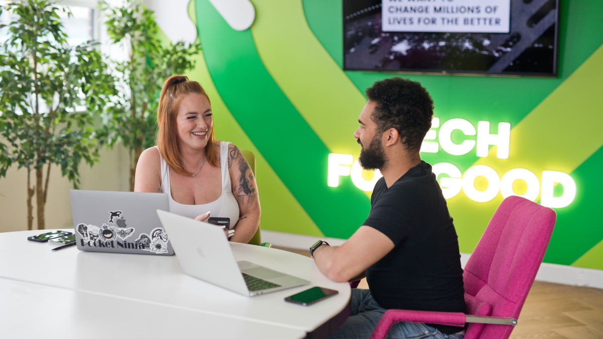 Two people smile and talk at a table with laptops. A bright "Tech For Good" sign is on the wall behind them.