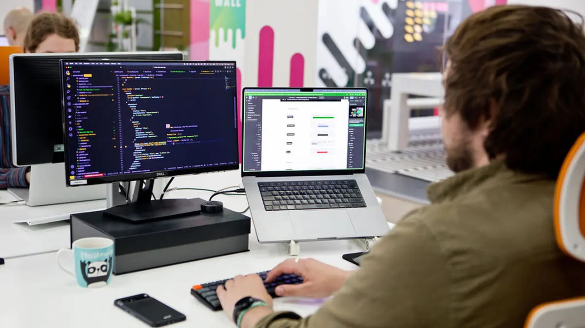 A person works at a desk with two monitors, one displaying code and the other a web design.
