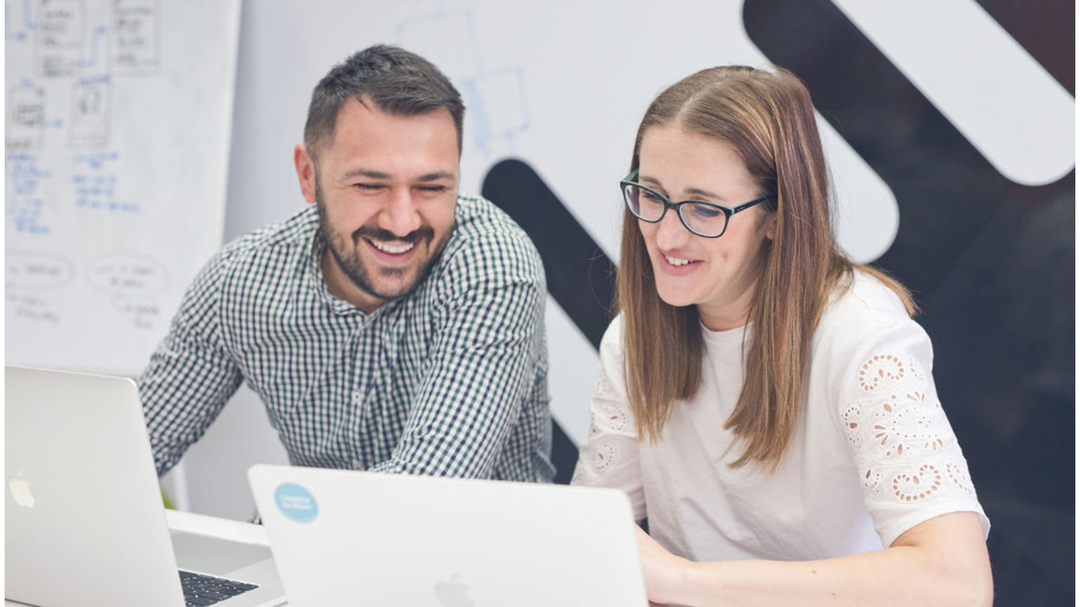 Two people smiling and working together on laptops, in a bright office setting with a whiteboard in the background.