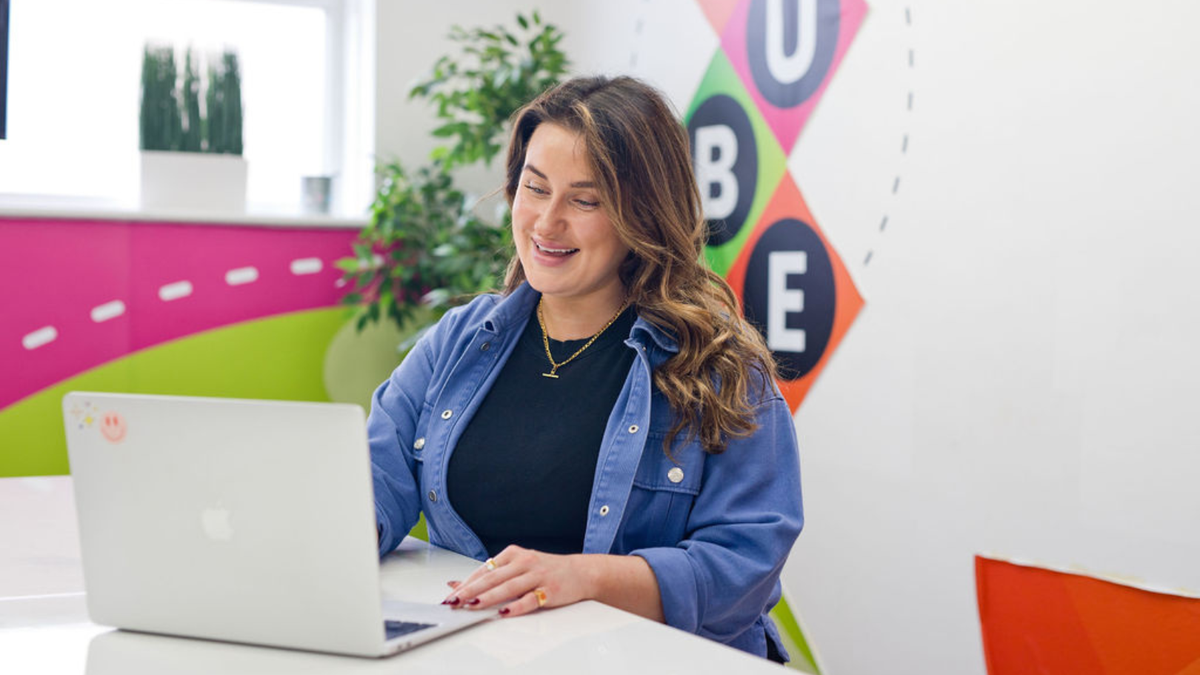Woman smiling while working on a laptop at a colorful office desk, surrounded by plants and a vibrant wall design.